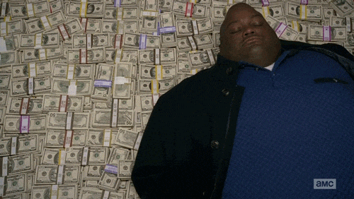 Huell, personaggio della serie tv Breaking Bad, si sdraia con un'espressione di puro compiacimento su una gigantesca montagna di mazzette di soldi all'interno di un magazzino